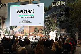 Bühne, Redner und Publikum bei einer Eröffnungsrede auf der Messe polis Convention 2021. Auf der Leinwand ist das Wort "Welcome" zu lesen.