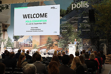 Bühne, Redner und Publikum bei einer Eröffnungsrede auf der Messe polis Convention 2021. Auf der Leinwand ist das Wort "Welcome" zu lesen.