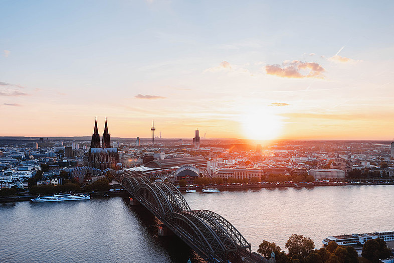 Blick von Deutz auf die Skyline der Stadt Köln mit dem Dom und dem Fernsehturm. bei Sonnenuntergang. Mittig erstreckt sich die Deutzer Brücke über den Rhein. 