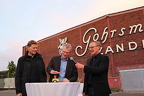 Bergisch Gladbachs Bürgermeister Frank Stein eröffnete in Anwesenheit von Dr. Reimar Molitor (Geschäftsführer REGIONALE 2025 Agentur) und Udo Krause (Leiter Projektgruppe Zanders) den Gohrsmühlenplatz und lässt den Schriftzug an der LFabrikhalle im Hintergrund per Knopfdruck aufleuchten.